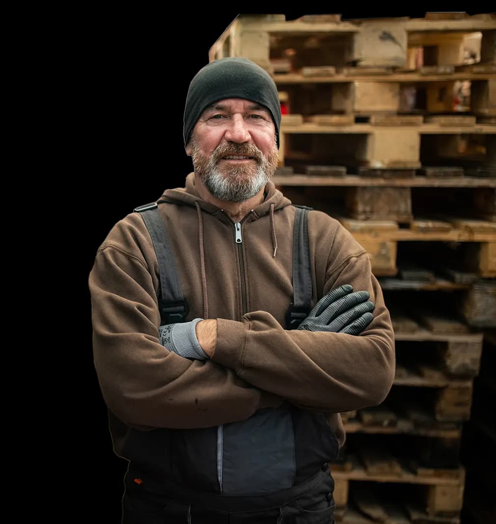 Man wearing sweater and beanie smiling and standing in front of a stack of pallets to haul-off