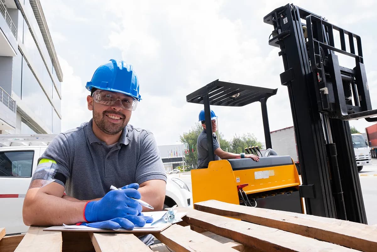 Smiling pallet yard worker in a hard hat and gloves inspecting wooden pallets, with a forklift behind him