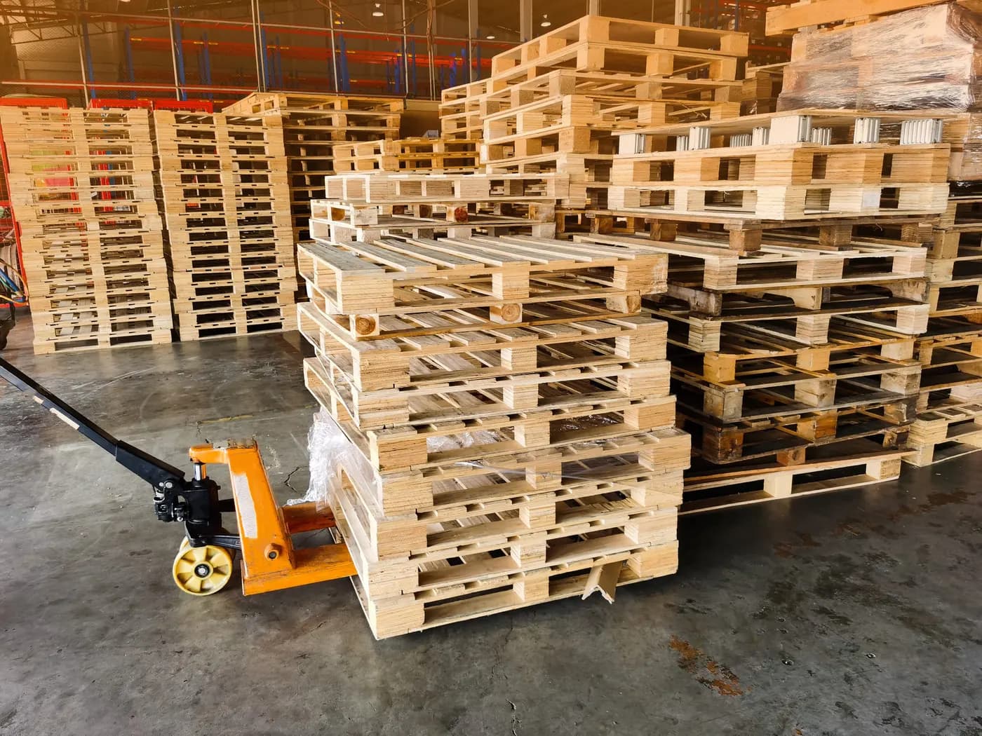 Stacks of wood pallets in a warehouse beside an orange pallet jack